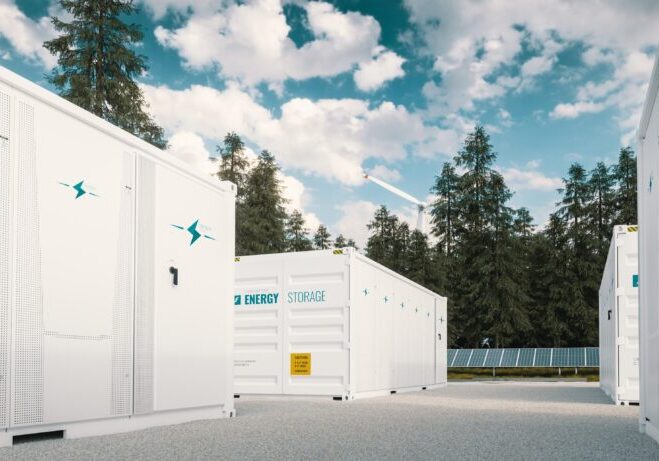 White energy storage containers are arranged outdoors on a gravel surface, surrounded by trees and a solar panel, with a partly cloudy sky overhead.